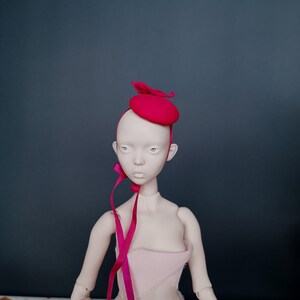 May include: A doll with a pale complexion wearing a fuchsia beret and matching ribbon. The beret has a leaf-like accent. The doll is dressed in a light pink garment against a dark teal background.