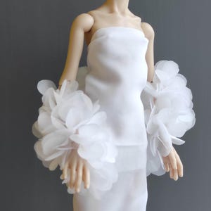 May include: A white strapless dress with matching floral arm cuffs. The cuffs are made of layered, sheer fabric petals. The dress is displayed on a doll against a grey background. The overall theme is bridal or formal wear.