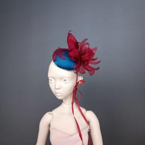May include: A teal blue fascinator with a large burgundy flower and a smaller embroidered detail. The headpiece is attached to a mannequin head with a burgundy ribbon. The background is a solid gray.