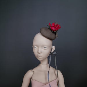 May include: A doll wearing a small, brown hat with a red flower accent. The hat is positioned on the doll's head, which is set against a dark gray background. The doll is wearing a pink dress and has gray ribbons tied around its ear.