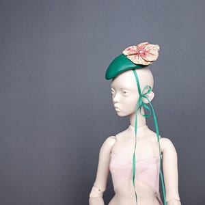 May include: A doll wearing a green leather beret with a pink and cream flower accent. The beret is adorned with long, teal ribbons tied in a bow. The doll is wearing a pink and white top.
