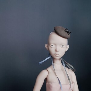 May include: A doll with a pale complexion and a small, brown hat decorated with a black bow. The doll has blue ribbons around its neck and is wearing a pink garment. The background is a solid, dark teal colour.
