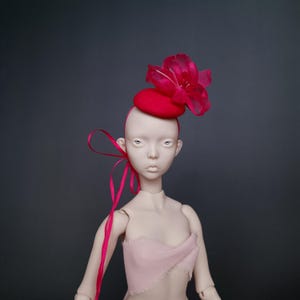 May include: A doll wearing a red hat with a large, sheer flower and a pink ribbon. The hat is a small, round design. The doll is wearing a pink top and has a neutral skin tone. The background is a solid, dark gray.
