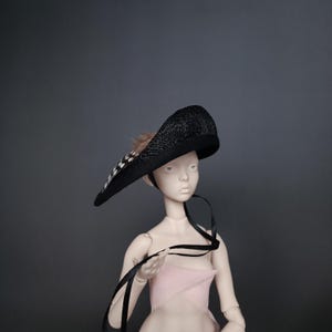 May include: A black hat with a wide brim, adorned with a feather accent. The hat is made of woven material and features a ribbon tie. The hat is on a doll with a pink dress.
