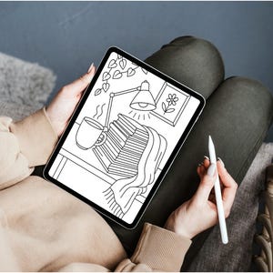 May include: A digital illustration on a tablet depicts a cozy scene with a stack of books, a steaming mug, and a desk lamp. A blanket drapes over the books. A stylus is held in the hand. The tablet is held on a person's lap.