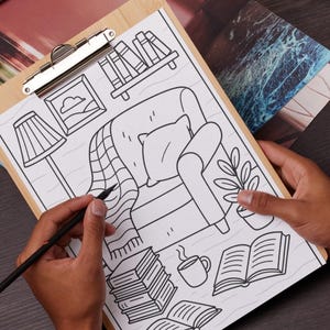 May include: A wooden clipboard holds a black and white illustration of a cozy living room scene. The drawing includes a chair, books, a lamp, and a plant. A hand holds a pencil, adding detail to the artwork.