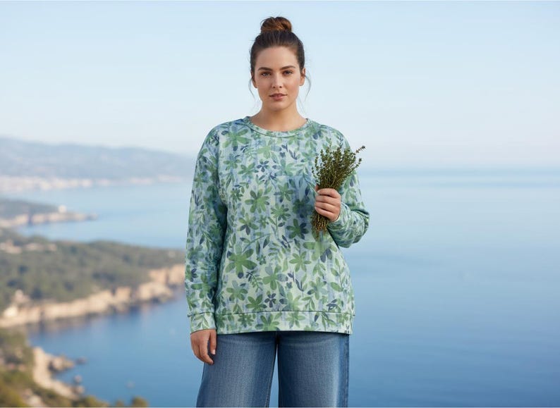 May include: A woman wearing a light blue sweatshirt with a floral pattern in shades of green and blue. She is holding a bundle of herbs. The background features a coastal landscape with a body of water and mountains.