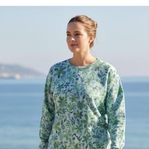 May include: A light blue and green floral print sweatshirt. The long-sleeved top features a crew neck and a pattern of green leaves and flowers. The background shows a blurred view of the ocean and sky.