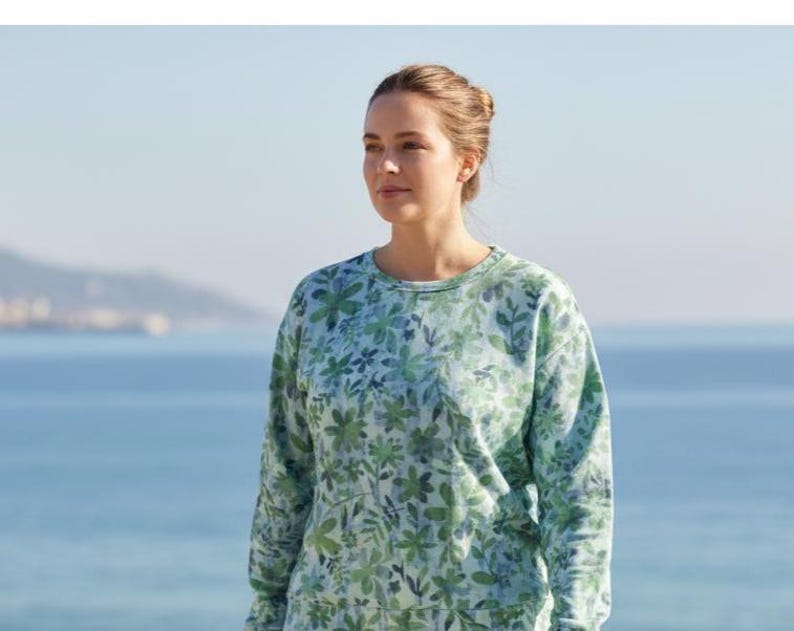 May include: A light blue and green floral print sweatshirt. The long-sleeved top features a crew neck and a pattern of green leaves and flowers. The background shows a blurred view of the ocean and sky.