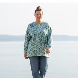 May include: A woman wearing a long-sleeved sweatshirt and blue jeans. The sweatshirt has a light blue background with a green floral pattern. The jeans are a medium wash denim.