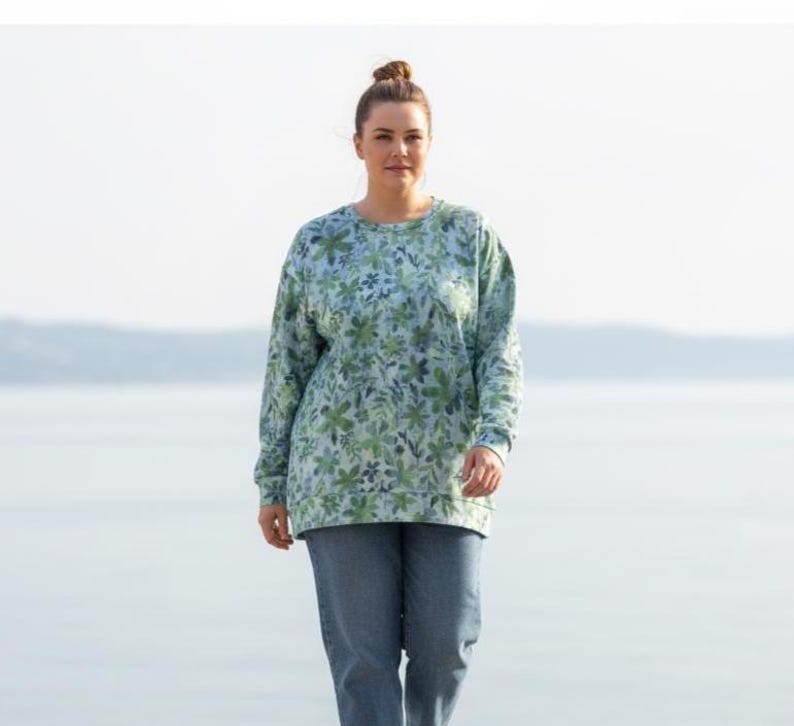 May include: A woman wearing a long-sleeved sweatshirt and blue jeans. The sweatshirt has a light blue background with a green floral pattern. The jeans are a medium wash denim.