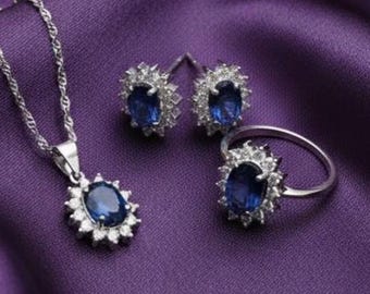 May include: A jewelry set featuring a silver necklace, earrings, and ring. Each piece has a central oval-shaped blue gemstone surrounded by smaller white stones. The set is displayed on a purple satin fabric.