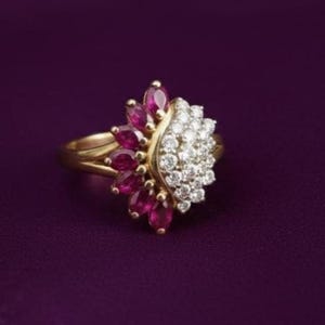 May include: A gold ring with a cluster of red and white gemstones. The ring features a floral design, with red stones on one side and white stones on the other. The ring is set against a deep purple background.