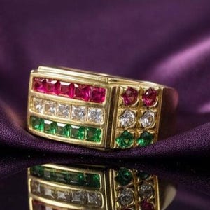 Könnte beinhalten: Ein goldener Ring mit rechteckiger Oberfläche, mit Reihen aus roten, weißen und grünen Edelsteinen. Der Ring hat eine polierte Oberfläche und ist vor einem violetten Hintergrund platziert, mit seiner Reflexion darunter.