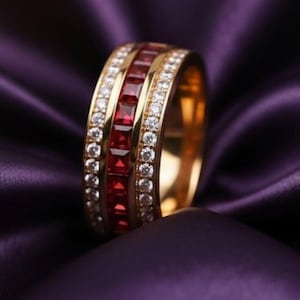 May include: A gold ring with a band of square-cut red gemstones and a row of small, clear gemstones. The ring is set against a backdrop of deep purple satin fabric.