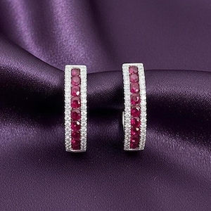 May include: A pair of rectangular hoop earrings. Each earring features a row of vibrant red gemstones set between rows of small, clear gemstones. The earrings are displayed on a purple satin fabric.