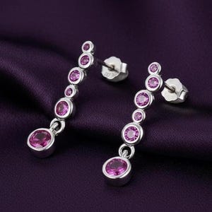 Puede incluir: Pendientes colgantes plateados con una serie de piedras preciosas rosas redondas y graduadas. Los pendientes tienen un cierre de poste y se muestran sobre un fondo morado oscuro.