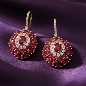 May include: A pair of gold drop earrings featuring a central oval-cut red gemstone surrounded by a halo of small white stones, with an outer layer of oval-cut red gemstones. The hook-style earrings have a pavé-set stone design on the wire.
