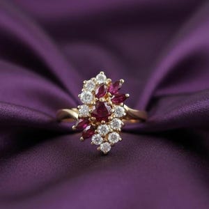May include: A gold ring with a cluster of gemstones. The ring features deep red rubies and sparkling white diamonds. The ring is set against a purple satin background, enhancing the jewellery's elegance and design.