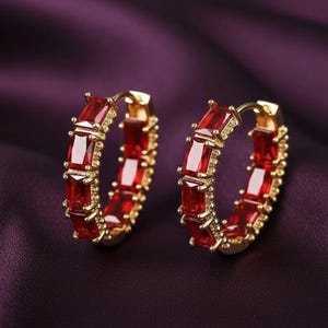 May include: A pair of gold hoop earrings featuring a series of rectangular, baguette-cut red gemstones set along the outer edge of each circular band. The vibrant red stones are held in place by small gold prongs, creating a continuous design.