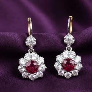 May include: A pair of elegant earrings featuring a central, round, deep red gemstone surrounded by a halo of smaller, clear gemstones. The earrings have a floral design and are set against a purple background.