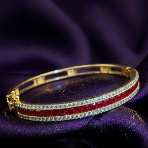 May include: Gold bangle bracelet featuring a central row of red gemstones, framed by smaller, light blue stones. The bracelet is displayed on a deep purple, textured fabric.