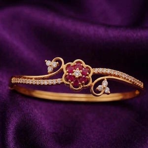 Pode incluir: Uma pulseira dourada com um design floral. A flor no centro é feita de pedras preciosas vermelhas, com uma única pedra preciosa branca no meio. A pulseira é adornada com pequenas pedras preciosas brancas.