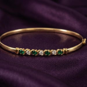 May include: A gold bangle bracelet with a row of alternating emerald and diamond gemstones. The bracelet is set against a dark purple background, highlighting the jewelry's sparkle and design.