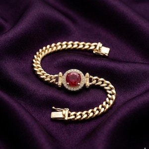 Peut inclure: Un bracelet en or avec une pierre centrale rouge entourée de petits diamants. Le bracelet présente un motif de chaîne gourmette et est posé sur un fond de satin violet foncé. Le fermoir est ouvert, révélant la construction du bracelet.