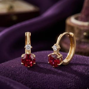 May include: A pair of gold hoop earrings featuring a round, deep red gemstone set in a six-prong basket, accented by a single small, clear, round-cut white stone positioned directly above the main red stone on the hoop mechanism.