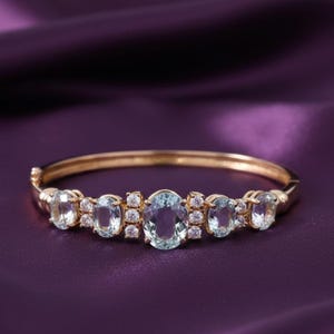 Könnte beinhalten: Ein goldfarbenes Armband mit ovalen, hellblauen Edelsteinen und kleinen, klaren Akzentsteinen. Das Armband ist vor einem tiefvioletten Hintergrund platziert, der das Design und die Farbe des Schmucks hervorhebt.