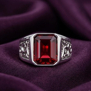 May include: A silver ring with a large, rectangular, deep red gemstone. The ring has ornate detailing on the sides and is set against a purple background. The gemstone is the focal point.
