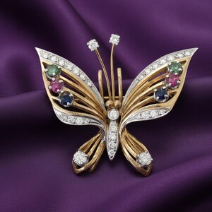 May include: A butterfly brooch crafted from gold and silver-toned metal, featuring clear round stones along the wings and body. Each upper wing is accented with a vertical row of three round-cut green, pink, and blue stones, plus two antennae.