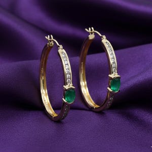 May include: A pair of gold hoop earrings with emerald green gemstones and small clear stones. The earrings are set against a purple background.