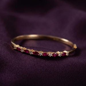 May include: A gold bangle bracelet featuring a row of alternating marquise-cut red rubies and round-cut clear white diamonds set along the front curve of the band, creating a delicate and symmetrical pattern of gemstones on the polished metal.