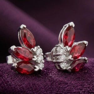 May include: A pair of silver stud earrings featuring three marquise-cut deep red gemstones arranged in a fan shape, accented by a vertical row of three small round clear stones set in a silver metal base.