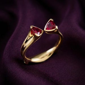 May include: A gold ring with two triangular, deep red gemstones. The ring's band curves gracefully, with each gemstone set in a gold bezel. The background is a rich, dark purple fabric, highlighting the ring's design.