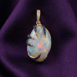 May include: A gold pendant with a large, oval opal gemstone. The opal displays iridescent colors and is accented with blue gemstones and gold claws. The pendant has a gold bail with small, clear gemstones. The background is a deep purple fabric.