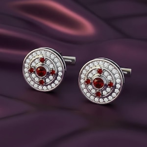 May include: Silver cufflinks with a circular design. Each cufflink has a central red gemstone, encircled by small white stones. Further red stones are arranged around the centre, forming a cross shape. The cufflinks are set against a purple background.