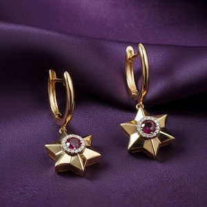 May include: A pair of gold-colored star-shaped earrings. Each earring features a central red gemstone surrounded by smaller white gemstones. The earrings hang from gold hoops. The background is a deep purple fabric.