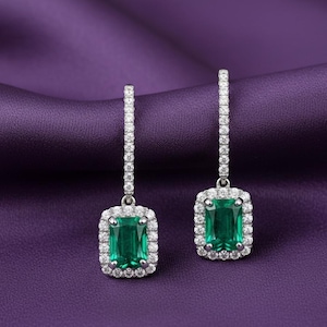 Puede incluir: Un par de pendientes colgantes de esmeralda y diamantes con corte esmeralda. Los pendientes presentan una piedra central de esmeralda verde vibrante de forma rectangular, rodeada por un halo de pequeños diamantes brillantes.