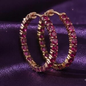 May include: A pair of hoop earrings crafted from gold-toned metal, each featuring a continuous row of round, vibrant pink gemstones set along the outer edge. The earrings have a hinged closure mechanism at the top for secure fastening.