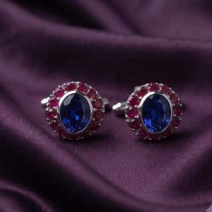 May include: A pair of silver-toned cufflinks featuring a central oval-cut deep blue gemstone surrounded by a halo of small, round, vibrant red stones. The metal settings are polished and circular, showcasing a classic design with a secure backing.