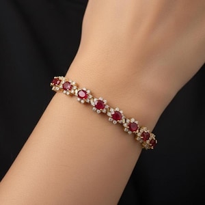 May include: A gold bracelet featuring oval-cut red gemstones and small, round, clear gemstones. The bracelet is set on a gold chain and is displayed on a wrist against a black background. The bracelet is a piece of jewelry.