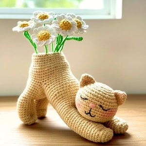May include: A beige crocheted cat-shaped vase holds white and yellow daisy flowers. The cat is lying down, with its head resting on its paws. The vase is a light tan colour and has a textured appearance.