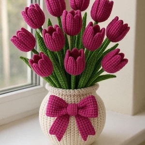 May include: A handcrafted crochet arrangement of pink tulips in a cream-colored vase with a matching bow. The tulips have green stems and leaves. The vase is sitting on a white windowsill.