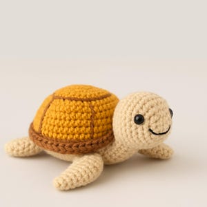 Puede incluir: Una tortuga de crochet hecha a mano. Tiene un caparazón amarillo mostaza con un borde marrón y una cabeza y extremidades beige. La tortuga tiene ojos negros y una boca sonriente. Un juguete suave para niños.