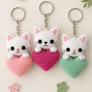 May include: Three handmade crochet cat keychains. Each white cat holds a heart in pink, fuchsia, and green. The cats have pink ears, black eyes, and a small pink nose. The keychains are attached to silver rings.