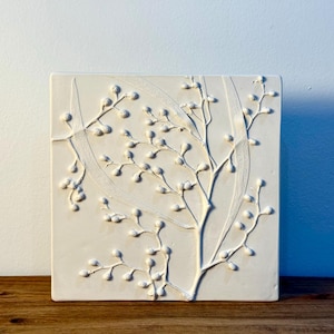 May include: A square, off-white ceramic tile with a raised relief design of a stylized tree branch with small, round buds. The artwork is set against a plain white wall and rests on a wooden surface.