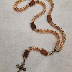 Puede incluir: Un rosario que presenta cuentas de vidrio craquelado translúcido de color ámbar intercaladas con cuentas marrones en forma de oso. El collar incluye una pieza central de metal color bronce y un crucifijo ornamentado a juego, unidos por un hilo dorado.